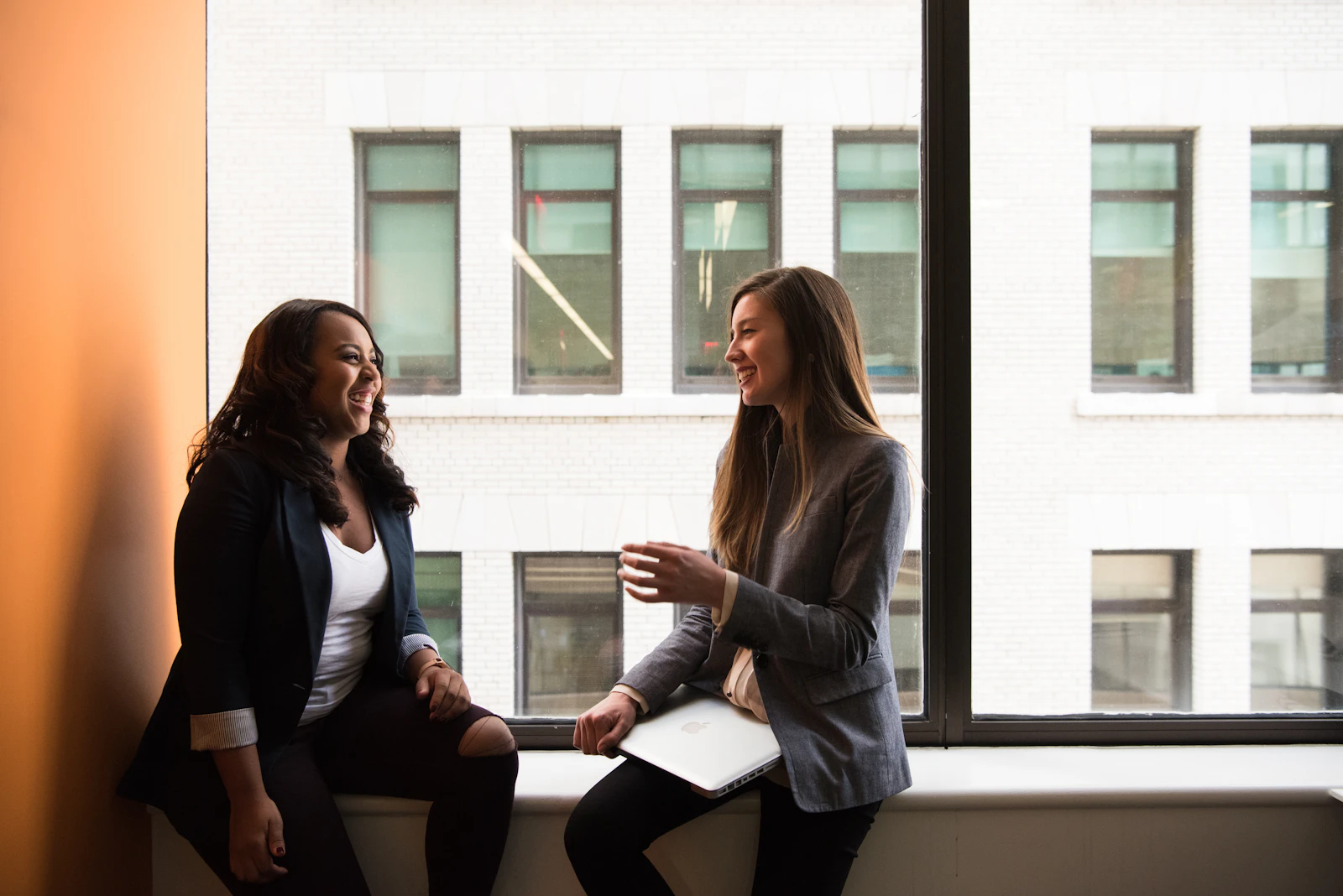 Deux collègues souriantes échangent près d’une fenêtre dans un espace de travail.